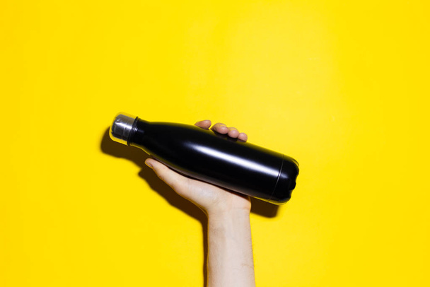 Close-up of male hand holding reusable steel thermo water bottle of black color. Isolated on yellow background. - Foto, Bild
