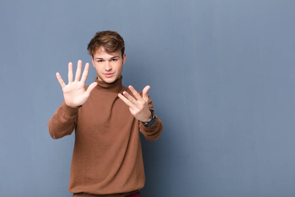 young blonde man smiling and looking friendly, showing number nine or ninth with hand forward, counting down isolated against flat wall - Foto, Imagen