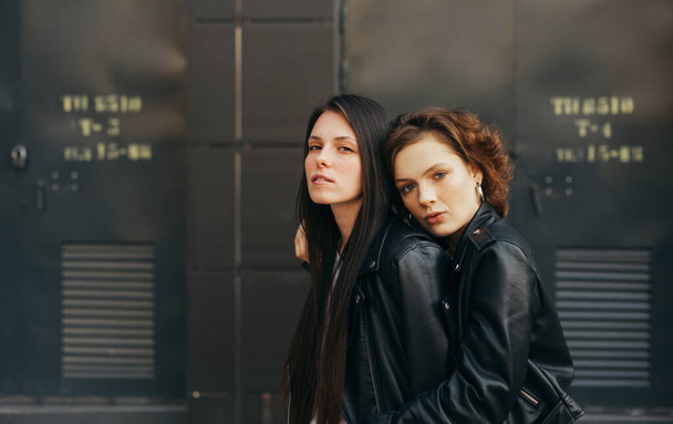 Detailní portrét dvou dívek v kožených bundách pózujících do kamery na pozadí tmavé stěny, dívajících se na kameru s vážnou tváří. Pár přítelkyň objímání na tmavém pozadí - Fotografie, Obrázek
