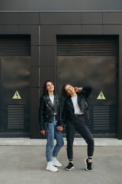 Vertical photo of two happy girlfriends having fun on black wall, looking at camera and smiling. Two cheerful girls are standing against a dark background and posing on camera. Walk with girlfriend. - Fotografie, Obrázek