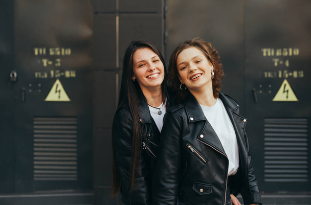 Couple filles heureuses en cuir foncé vestes debout contre un mur noir, en regardant la caméra et en souriant sincèrement. Deux copines se tiennent debout sur un fond sombre et sourient
 - Photo, image
