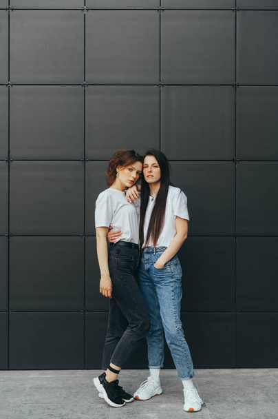 Vertical photo. Beautiful girls in casuals cuddling on black background and looking at the camera. Couple of beautiful girlfriends are standing on black background and posing at camera. Copy space - Фото, изображение