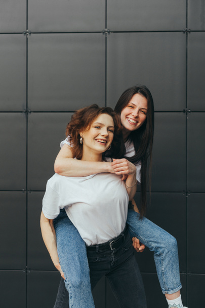 Portrait of cheerful two girlfriends having fun on dark background, girl carry girlfriend on her back, piggy back, looking at camera and smiling. Happy 2 girls having fun on black wall background - Foto, imagen