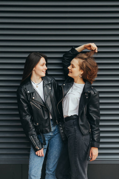 Happy couple girls in casual casual clothes are standing against a black wall background, talking and smiling, wearing leather jackets.Two cheerful girlfriends laughing on dark background. Vertical - Foto, afbeelding