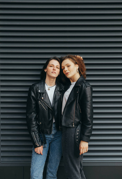 Fashionable photo of 2 girls in casual clothes standing against a dark background and looking in camera with a serious face wearing leather jackets. Attractive brunette girl on a black wall background - Photo, Image