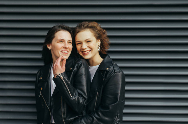 Closeup portrait of a couple of happy girls on black background, talking and smiling, wearing dark casual clothes. Smiling girlfriends in leather jackets stand against a dark background and have fun - Photo, Image