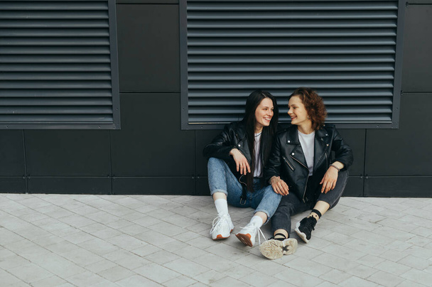 Twee gelukkige meisjes in stijlvolle casual kleding zitten op een stoep tegen een donkere muur achtergrond, kijken elkaar aan en glimlachen.Straatfoto van twee vriendinnen die op de grond liggen - Foto, afbeelding