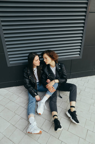 Modieuze foto van twee meisjes in leren jassen en jeans zittend op een ondergrond tegen een donkere muurachtergrond. Vriendinnen gingen zitten voor een pauze op de wandeling, meisje kijkt naar vriendin - Foto, afbeelding