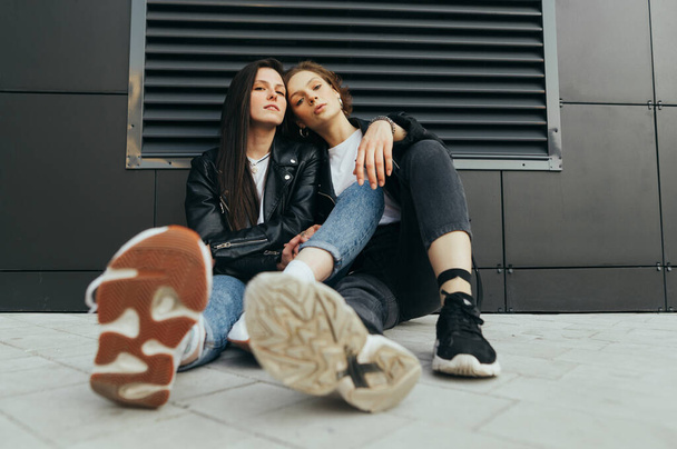 Fashionable photo of two stylish girls in casual clothes wearing leather jackets and sneakers,sitting on the ground against a dark wall and posing in camera.Fashion portrait 2 girlfriends on street - Фото, изображение