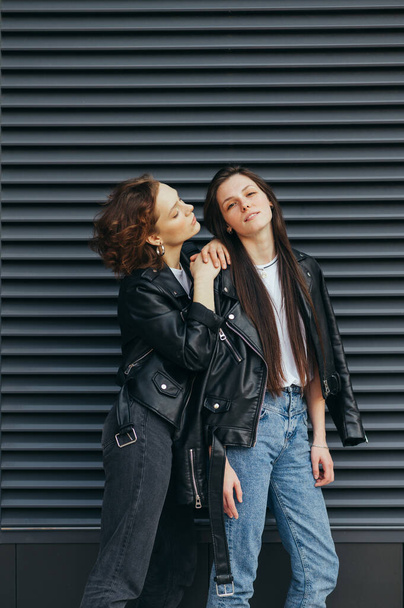 Vertical photo of two girlfriends in leather jackets posing on camera against a dark wall background. Fashionable girl in casual clothes stand on black background. Copy space - Photo, Image