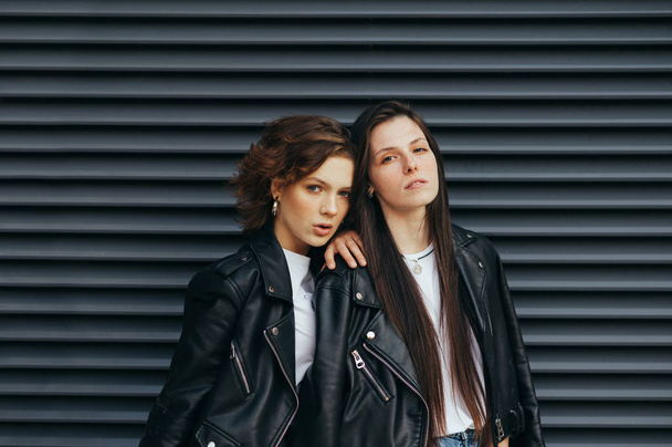 Modieus portret van twee modellen met sproeten in casual kleding op een donkere straat achtergrond, het dragen van leren jassen, kijken in de camera met een serieus gezicht.Meisje poseren op de camera op donkere muur - Foto, afbeelding