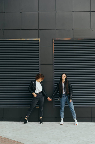 Vertical photo of two stylish girls in casual clothes on black background holding hands and posing at camera. Minimalistic photo, two girlfriends standing against a dark wall and looking at camera - Foto, afbeelding