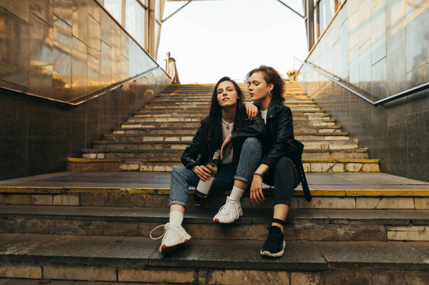 Photo à la mode de deux filles en vêtements décontractés élégants assis sur les escaliers à la sortie du métro, tenant une bouteille de vin et posant à la caméra.
 - Photo, image