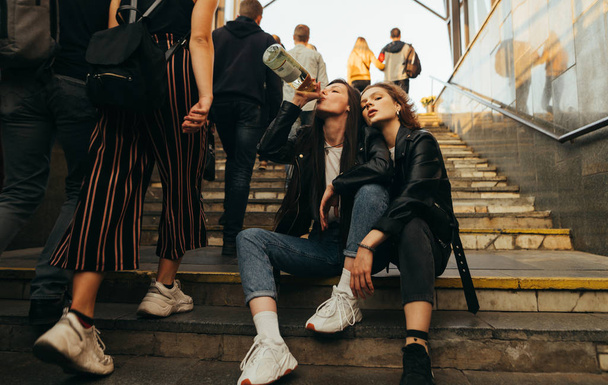Freedom girls sit on the stairs in a crowd of people and drink wine. Girlfriends drinking alcohol from a bottle sitting on the stairs at the exit of the subway on a background of passersby. - Foto, imagen