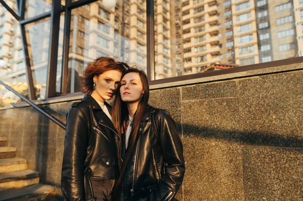 Straatportret van twee aantrekkelijke meisjes in leren jassen die op de achtergrond van een gebouw staan en met een serieus gezicht voor de camera poseren. Portret van aantrekkelijke vriendinnen in warme kleuren. - Foto, afbeelding
