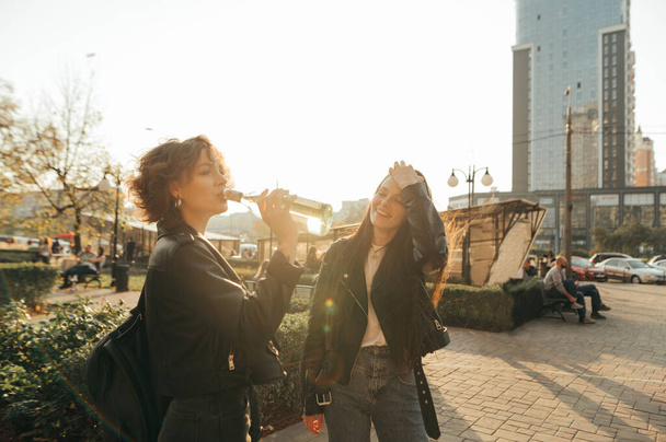 Happy two girls in casual clothes are standing outside at sunrise and drinking wine from bottles, smiling on cityscape background and wearing leather jackets. Freedom girls have fun on a walk. - Photo, Image