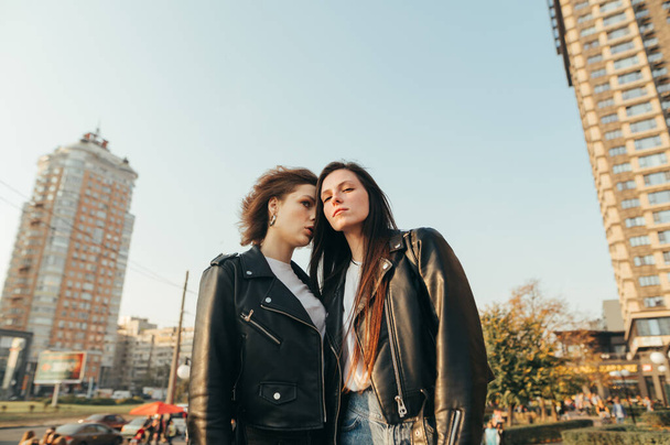 Fashion portrait of two girls in street clothes standing on grass on cityscape background and posing at camera. Portrait of attractive models in leather jackets on the street. - Photo, Image