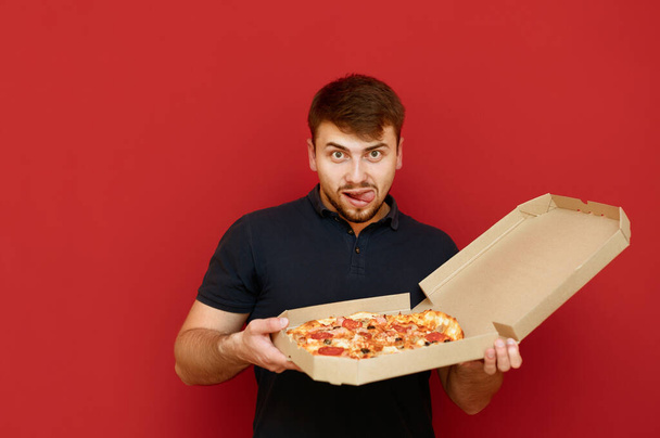 Zabawny facet z brodą trzyma pudełko po pizzy i patrzy w kamerę z głodną pożądliwą twarzą. Portret głodnego mężczyzny z pudełkiem po pizzy na czerwonym tle. Koncepcja fast-foodów. - Zdjęcie, obraz