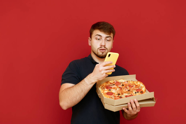 A young man with a beard holds a box of fresh pizza and uses a smartphone on a red background, looking intently at the Internet. Pizza delivery concept. Customer received the pizza delivered - Photo, Image