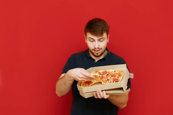 Homem engraçado com fome fica em um fundo vermelho e tira uma fatia de pizza da caixa. Um jovem engraçado com barba a comer pizza fresca da caixa. Isolados
. - Foto, Imagem