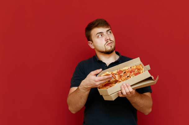 Man with a box of pizza stands on a red background, takes a slice of fast food and looks away with a serious face. Hungry bearded man eating hot pizza. Isolated. Copy space - Foto, Bild