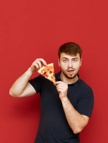 Retrato de un hombre guapo con una barba de pie sobre un fondo rojo con un pedazo de pizza en las manos, mira a la cámara y sonríe. Hombre posando en cámara con rebanada de pizza, aislado. Copiar espacio
 - Foto, Imagen