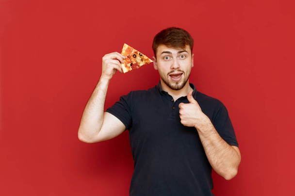 Positive man with beard stands on red background with piece of pizza in hand, looks into camera, smiles and shows thumb up. Hungry guy poses with a slice of pizza and shows a gesture of liking - Foto, afbeelding