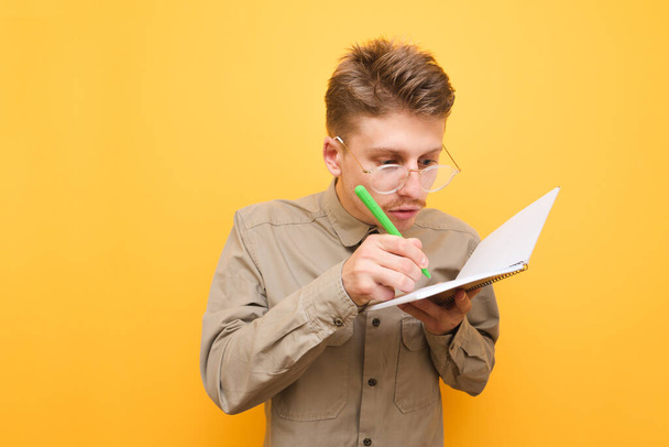 Funny geek wearing glasses and a shirt stands on a yellow background and writes in a notebook in a focused manner. Funny nerd with mustache notes in a notebook. Student doing homework - Photo, Image