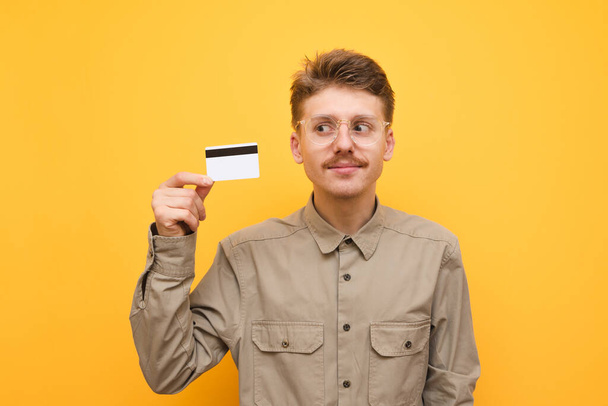 Close-up portret van een grappige nerd met een bankkaart in zijn hand op een gele achtergrond, wegkijkend en glimlachend. De man in het shirt en de bril heeft een creditcard in zijn hand. Geïsoleerd. Kopieerruimte - Foto, afbeelding