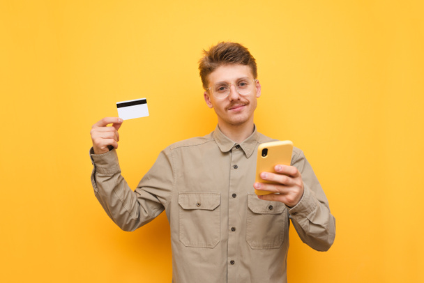 Positive young man with glasses and mustache holding credit card and smartphone, looking into camera and smiling against yellow background. Cheerful guy makes purchases online and poses for the camera - Photo, Image