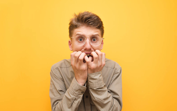 Portrait of a scared young man in shirt and glasses on yellow background, looks into camera with shocked face and bites his fingers. Scared nerd isolated on yellow background. Copy space - Foto, Bild