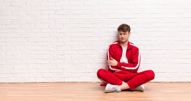 young blonde man feeling sad, upset or angry and looking to the side with a negative attitude, frowning in disagreement sitting on the floor - Photo, Image