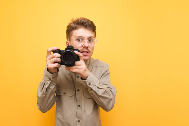 Portrait of funny stock photographer with glasses and shirt on yellow background,takes photo on camera and with serious face looks into camera.Nerd photographer with camera in hands isolated on yellow - Photo, Image