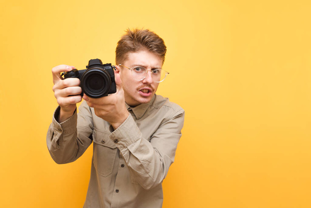 Portrait of funny paparazzi photographer with glasses and wears shirt on yellow background, holds camera in hand and makes photo, looks into camera with serious face. Copy space - Fotografie, Obrázek