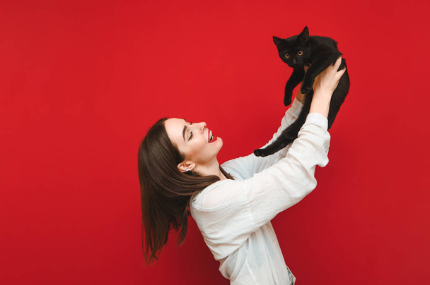 Portrait de fille heureuse sur fond rouge jouant avec chat dans les mains, souriant tout en regardant l'animal de compagnie. Fille souriante en chemise blanche jouant avec chat noir sur fond, isolé. Espace de copie
 - Photo, image