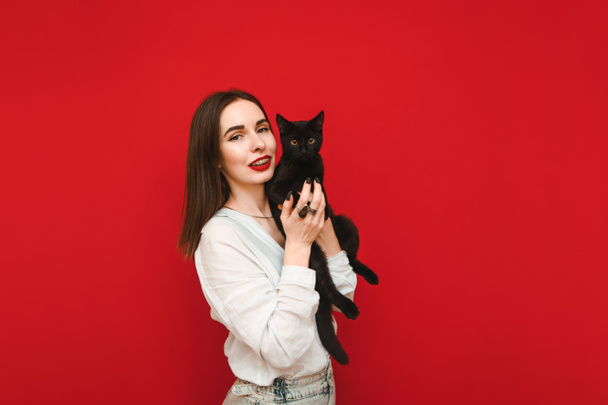 Uśmiechnięta kobieta w lekkich, luźnych ubraniach stoi na czerwonym tle z czarnym kotem w rękach, patrzy w kamerę i uśmiecha się. Szczęśliwy czarny kot właściciel odizolowany na czerwonym tle. Pojęcie zwierząt domowych. - Zdjęcie, obraz
