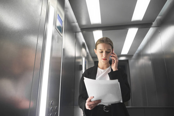 Serious businesswoman is talking by phone looking at papers in the elevator. Young female employee is reading the documents and talking by smartphone in the lift. - Foto, Imagem