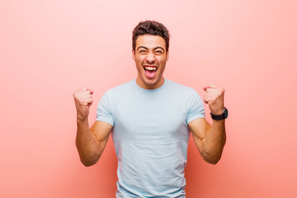 young arabian man shouting aggressively with an angry expression or with fists clenched celebrating success against pink wall - Photo, Image
