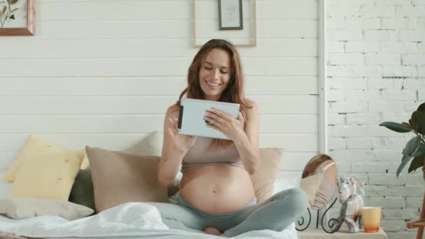 Šťastná těhotná žena hledá tablet v posteli. Belly matka drží ipad doma. - Záběry, video