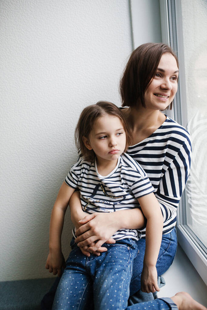 Happy young mother with her upset girl child at home near window, short brunette hair, same clothes, woman smiling  - Foto, imagen