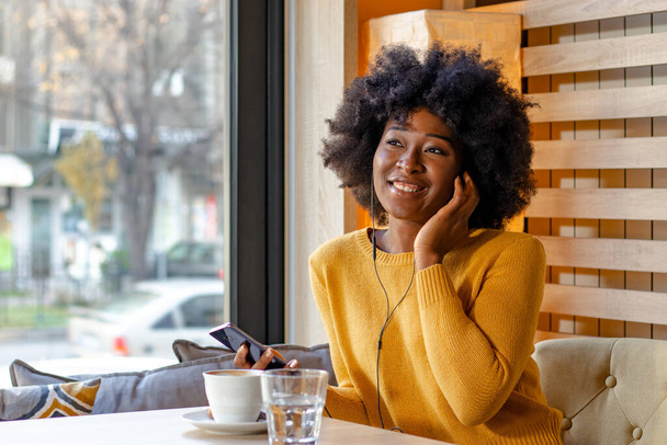 Black african beautiful young woman, sitting in cafe, listening to music or talking via mobile phone with earbuds in her ears and enjoying a cup of coffee. - Φωτογραφία, εικόνα