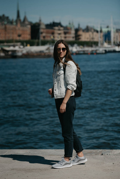 A young female tourist in a white jeans jacket, black jeans, grey sneakers, sunglasses, and backpack waits on the center of the European town near the water - 写真・画像