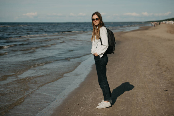 A young female tourist in a white jeans jacket, black jeans, grey sneakers, sunglasses, and backpack poses near the sea - Fotografie, Obrázek