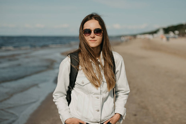 Una joven con chaqueta blanca, jeans negros, zapatillas grises, gafas de sol y mochila posa cerca del mar
 - Foto, imagen