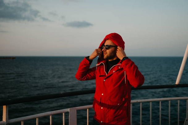 赤いジャケットとサングラスをしたスタイリッシュな男は、夕日を見て船に乗って頭の上にフードを着ます。背景にドライ貨物船. - 写真・画像