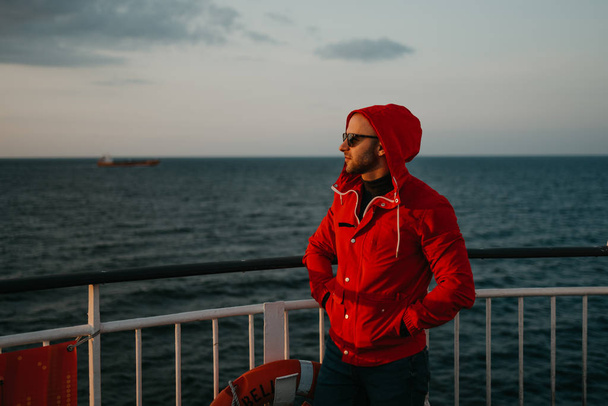 フードの赤いジャケットとサングラスに身を包んだスタイリッシュな男は、夕日を見て船に乗って頭の上にフードをかぶっています。背景にドライ貨物船. - 写真・画像