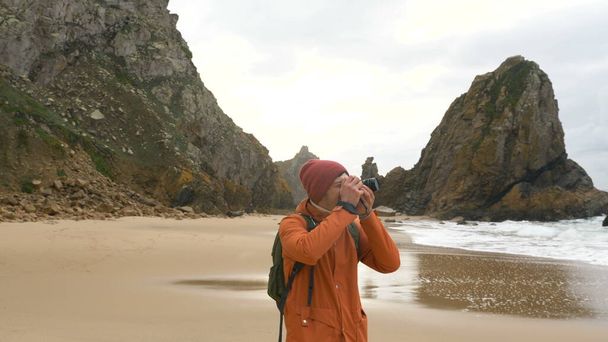 A traveler takes pictures with a modern camera of landscapes on the ocean shore among rocks and large waves - Photo, Image