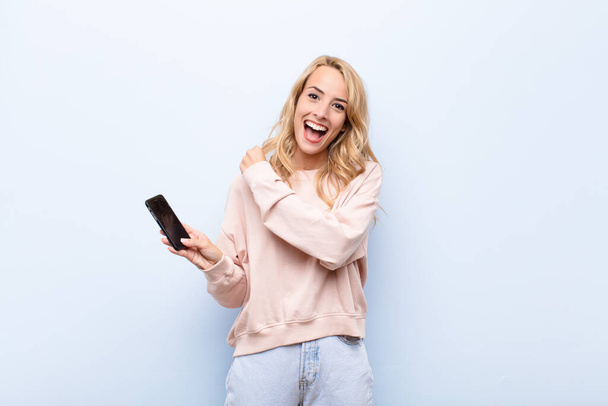 young blonde woman feeling happy, positive and successful, motivated when facing a challenge or celebrating good results using a smartphone - Valokuva, kuva