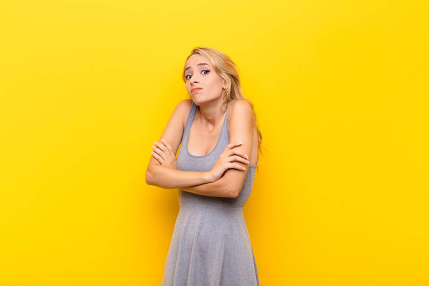 young blonde woman shrugging, feeling confused and uncertain, doubting with arms crossed and puzzled look against orange wall - Photo, image