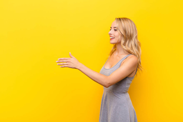 young blonde woman smiling, greeting you and offering a hand shake to close a successful deal, cooperation concept against orange wall - Foto, Imagem
